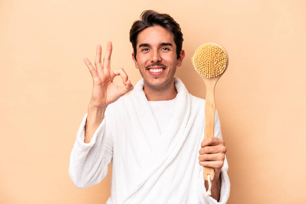 Young caucasian man wearing a bathrobe holding a back scratcher isolated on beige background cheerful and confident showing ok gesture. - Foto, afbeelding