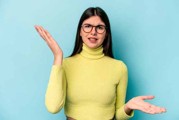 Young caucasian woman isolated on blue background confused and doubtful shrugging shoulders to hold a copy space. - Photo, Image