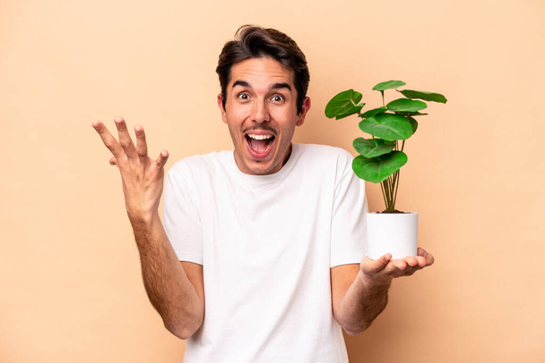 Fiatal kaukázusi férfi kezében egy növény elszigetelt bézs háttérrel kap egy kellemes meglepetés, izgatott és felemelt kezét. - Fotó, kép