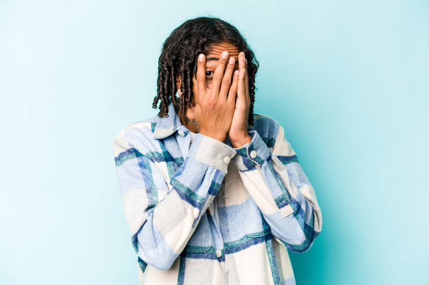 Νεαρός Αφροαμερικάνος απομονωμένος σε μπλε φόντο αναβοσβήνει μέσα από τα δάχτυλα φοβισμένος και νευρικός. - Φωτογραφία, εικόνα