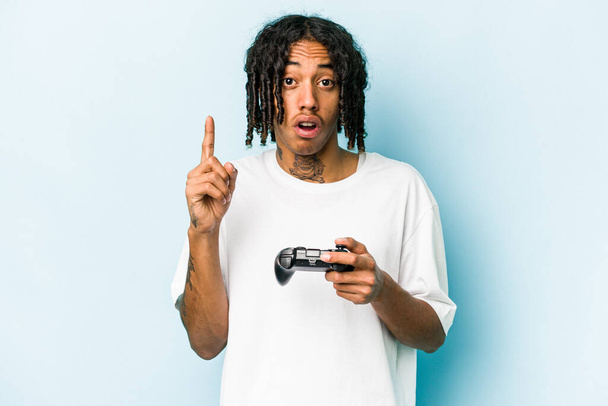 Young African American man playing with a video game controller isolated on blue background having some great idea, concept of creativity. - Zdjęcie, obraz