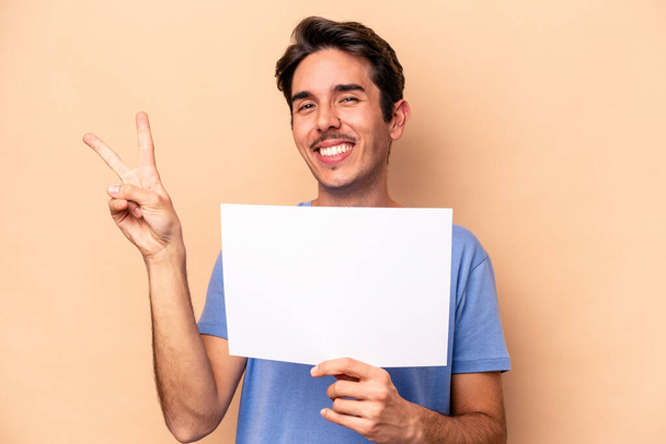 Young caucasian man holding a placard isolated on beige background joyful and carefree showing a peace symbol with fingers. - Photo, Image