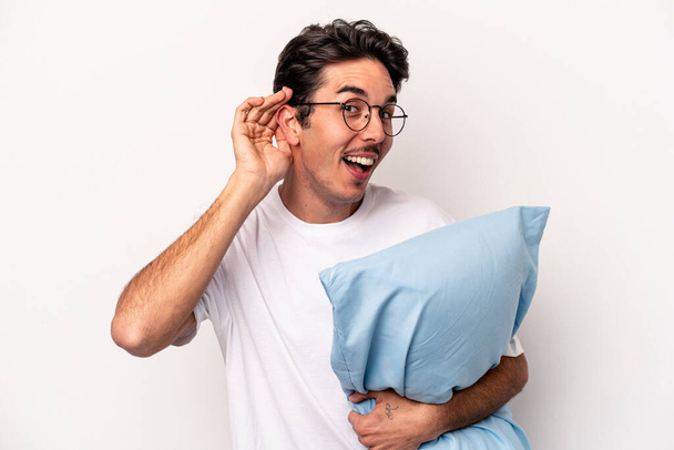 Junger kaukasischer Mann im Pyjama hält ein Kissen isoliert auf weißem Hintergrund und versucht, einem Klatsch und Tratsch zuzuhören. - Foto, Bild