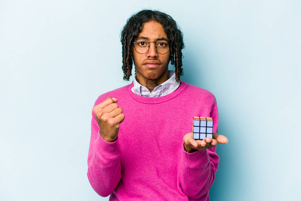 Junger afroamerikanischer Mann hält einen Rubiks-Würfel isoliert auf blauem Hintergrund und zeigt der Kamera die Faust, aggressiver Gesichtsausdruck. - Foto, Bild