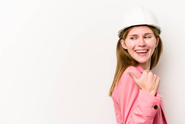 Young architect English woman with helmet isolated on white background points with thumb finger away, laughing and carefree. - Photo, Image