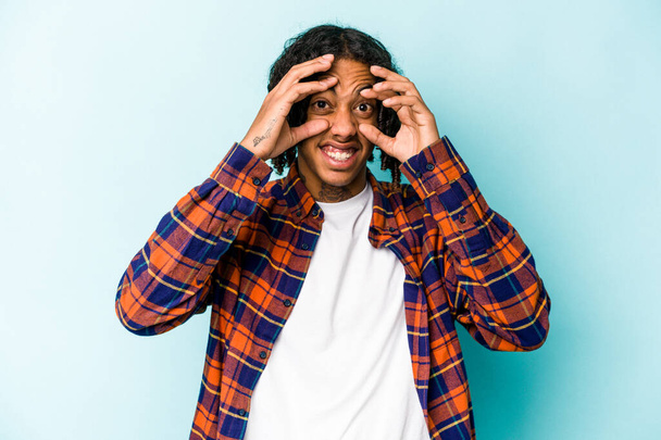 Νεαρός Αφροαμερικάνος απομονωμένος σε μπλε φόντο κρατώντας τα μάτια ανοιχτά για να βρει μια ευκαιρία επιτυχίας. - Φωτογραφία, εικόνα