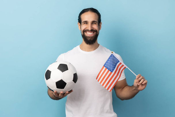 Portrait of satisfied delighted man wearing white T-shirt holding flag of united states of america and soccer black and white ball, united soccer league. Indoor studio shot isolated on blue background - Фото, изображение