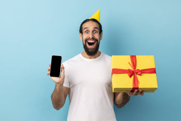 Portrait of man wearing white T-shirt in birthday cone holding gift box and cell phone with mockup, copy space for advertisement, blank display. Indoor studio shot isolated on blue background. - Foto, afbeelding
