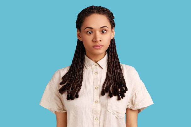 Portrait of funny silly woman with black dreadlocks looking at camera, cross eyed with stupid dumb face, has awkward confused comical expression. Indoor studio shot isolated on blue background. - Fotografie, Obrázek