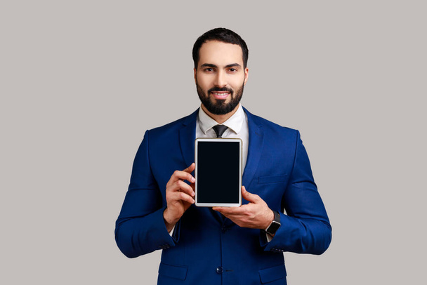 Man holding and showing tablet with blank screen with toothy smiling and satisfied face, copy space for advertisement, wearing official style suit. Indoor studio shot isolated on gray background. - Zdjęcie, obraz