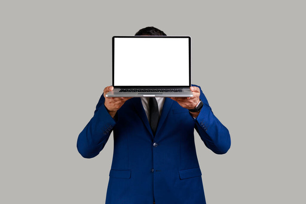 Anonymous unknown man hiding his face behind laptop with white blank display with space for advertisement, wearing official style suit. Indoor studio shot isolated on gray background. - Фото, зображення