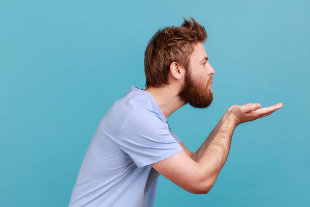 Side view of bearded man blowing air kiss keeps lips, folded palm towards mouth, sends mwah coquettish, wants to cheer up her partner. Indoor studio shot isolated on blue background. - Foto, imagen