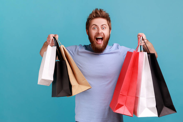 Portrait of amazed excited handsome bearded man holding shopping paper bags, pleased and happy with mall discounts, good purchases. Indoor studio shot isolated on blue background. - Foto, Imagem