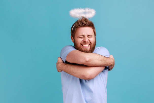 Portrait of bearded angelic man with holy nimbus standing smiling contentedly and hugging himself, self-love, support, keeps eyes closed. Indoor studio shot isolated on blue background. - Photo, image