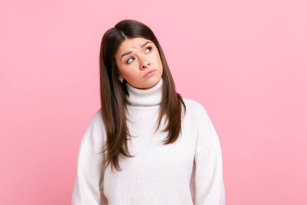 Confused puzzles female looking thoughtfully away with pensive expression, making decision, solving dilemma, wearing white casual style sweater. Indoor studio shot isolated on pink background. - Foto, imagen