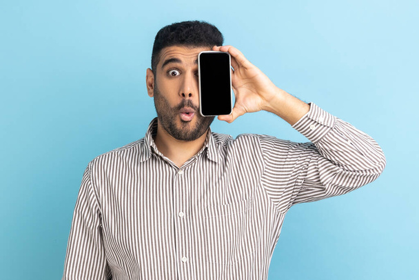 Portrait of shocked handsome businessman standing, holding and covering one eye with smartphone, looking at camera, wearing striped shirt. Indoor studio shot isolated on blue background. - Photo, image