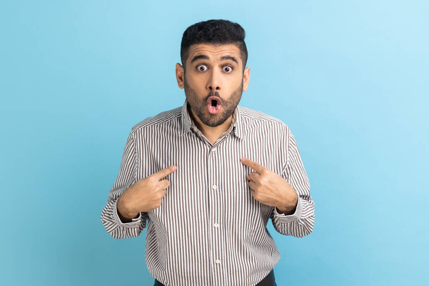 Businessman pointing finger on itself looking at camera with surprised expression, wondered with sudden victory, hear unbelievable news, wearing shirt. Indoor studio shot isolated on blue background. - Foto, Imagem