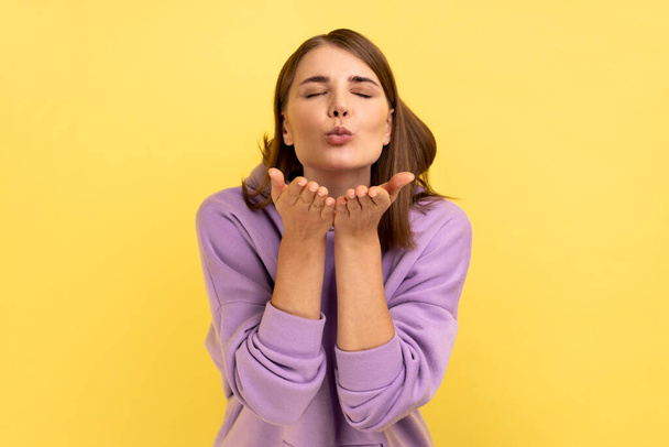 Woman sending air kiss to camera, flirting and demonstrating love affection feelings, keeps eyes closed, romantic relationships, wearing purple hoodie. Indoor studio shot isolated on yellow background - Valokuva, kuva