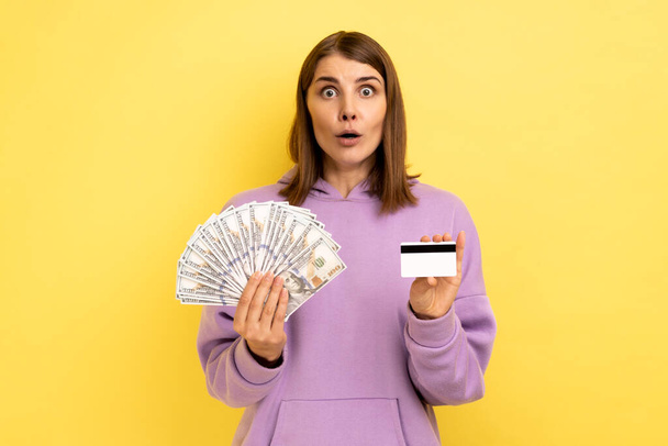 Portrait of shocked woman with dark hair holding fan of dollar banknotes and credit card, earning money, banking, wearing purple hoodie. Indoor studio shot isolated on yellow background. - Fotografie, Obrázek