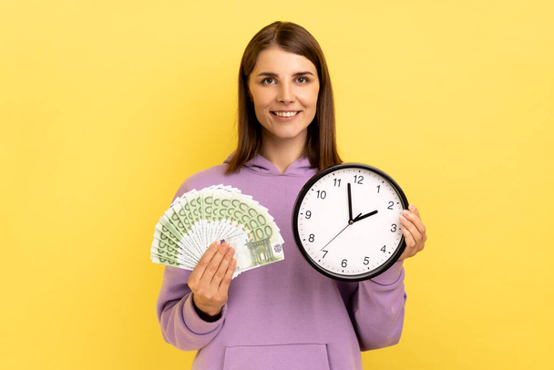 Portrait of rich dark haired woman holding big fan of euro banknotes and wall clock, time is money, expressing happiness, wearing purple hoodie. Indoor studio shot isolated on yellow background. - Foto, Bild