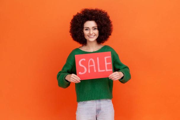 Smiling woman with Afro hairstyle wearing green casual style sweater standing holding card with sale inscription, presenting huge discounts. Indoor studio shot isolated on orange background. - Foto, Bild
