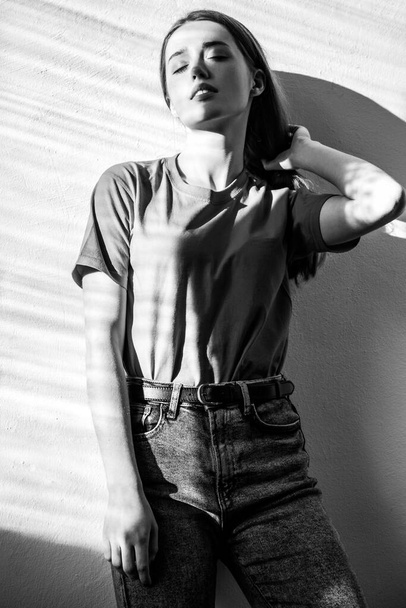 Beautiful woman wearing T-shirt and jeans standing with dreamy romantic expression, keeps eyes closed. Black and white photography, indoor studio shot illuminated by sunlight from window - Foto, Imagem