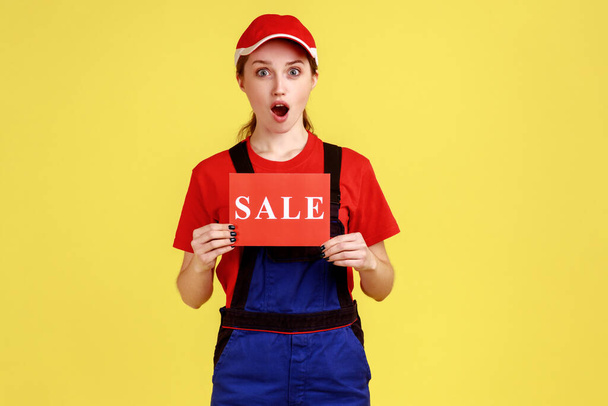Portrait of amazed shocked handy woman standing and holding card with sale inscription, discounts for service, wearing overalls and red cap. Indoor studio shot isolated on yellow background. - Фото, изображение