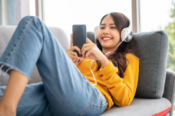Bella asiatica giovane donna che indossa auricolari e utilizzando smartphone mobile. Tempo libero femminile sul divano e godere di guardare lo schermo del dispositivo smartphone - Foto, immagini