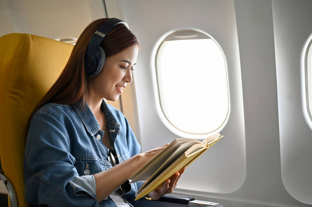 Расслабленная азиатка-путешественница тысячелетия в самолёте, слушающая музыку в беспроводных наушниках и читающая книгу на своём месте. Концепция путешествия - Фото, изображение