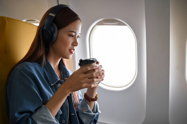 Kulaklıklı çekici Asyalı kadın uçakta sıcak bir kahve yudumlarken rahatlıyor. Seyahat, tatil, turizm konsepti - Fotoğraf, Görsel