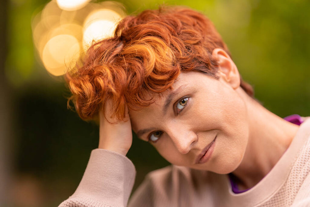 Glad androgynous woman touching short red hair and looking at camera while spending summer day in park - Fotografie, Obrázek