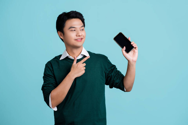 Портрет красивого азиатского мужчины, показывающего или представляющего мобильное приложение на синем фоне. - Фото, изображение