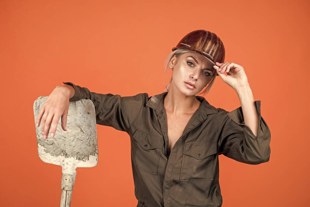 woman worker in protective helmet and boilersuit hold shovel on orange background, labouring. - Foto, Bild
