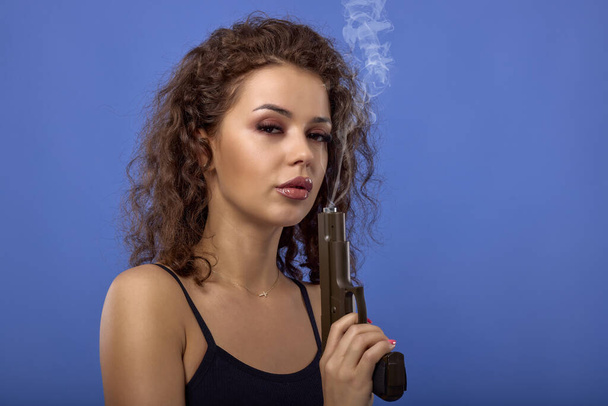 beautiful young woman with curly hair with a gun on a blue background - Fotó, kép