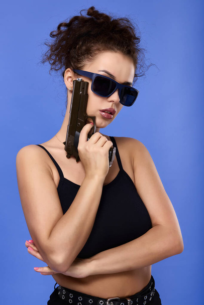 beautiful young woman with curly hair with a gun on a blue background - 写真・画像