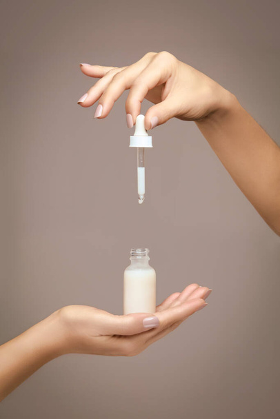 Female hands holding an unlabelled generic dropper bottle filled with white cosmetic fluid over a beige background in a cosmetology and beauty concept - Fotografie, Obrázek
