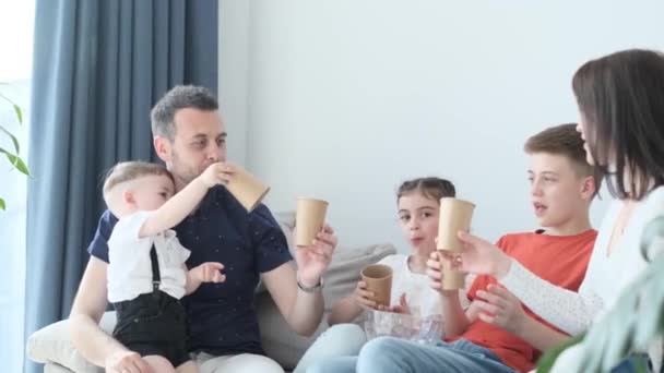 Όμορφη ευρωπαϊκή οικογένεια χαλαρώνοντας στο σπίτι μαζί στον καναπέ, γιορτάζουν τα γενέθλιά τους και πίνουν από χάρτινα ποτήρια. Ευτυχισμένη οικογένεια στο σπίτι - Πλάνα, βίντεο