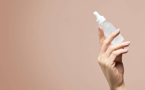 Weibliche Hand hält eine undurchsichtige, unetikettierte Tropfflasche in Kosmetik- und Hautpflegekonzept hoch, mit seitlichem Kopierraum für Ihren Text auf beigem Hintergrund - Foto, Bild