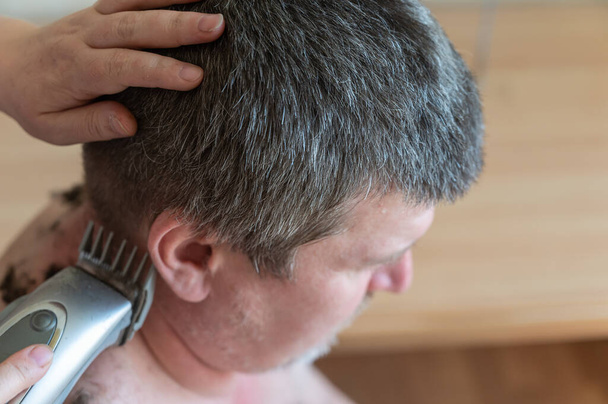 Wife cuts her husband's hair at home. Woman uses an electric hair clipper to cut man's hair at back of his head. Middle-aged person with naked torso sits inside living room. Selective Focus. - Photo, Image
