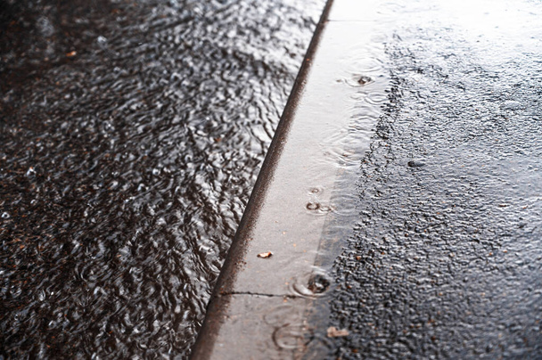 Niederschlag auf einer Stadtstraße entlang des Bordsteins. Sommergewitter. Wasserstrahl aus einem Regenguss. Fallende große Regentropfen. Selektiver Fokus. Bewegung verschwimmt. Lärm, Getreideeffekt. - Foto, Bild