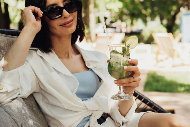 Young Brunette Woman Relaxing and Drinking Cocktail While Sitting on the Restaurant Terrace Outdoors in Summer Sunny Day - Photo, image