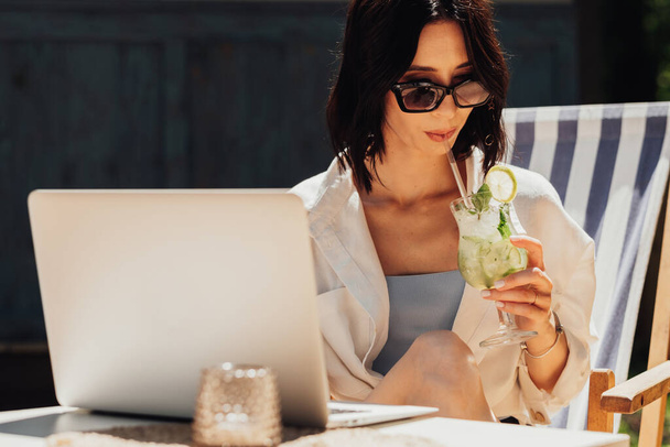 Jovem morena de óculos de sol trabalhando no laptop e bebendo coquetel enquanto sentado no terraço do restaurante, freelancer no trabalho - Foto, Imagem