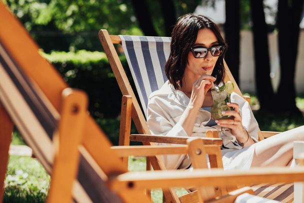 Güneş gözlüklü genç esmer kadın terasta otururken kokteyl içiyor kız güneşli yaz gününün tadını çıkarıyor. - Fotoğraf, Görsel