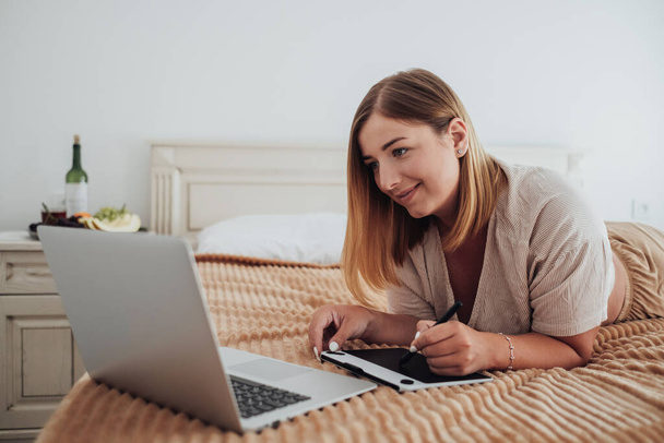 Vrolijke blanke vrouw op bed in het hotel kamer en werken aan een digitale tablet en laptop, Ontwerper Freelancer op het werk - Foto, afbeelding