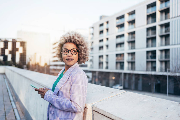 Professionele zwarte zakenvrouw met blond haar en trendy brillen die in de stad staan en weg kijken bij zonsondergang - Foto, afbeelding