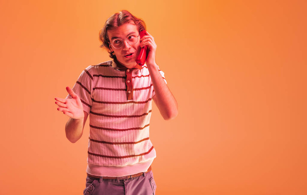 Porträt eines emotionalen jungen Mannes, der mit einem Retro-Telefon telefoniert und isoliert vor orangefarbenem Studiohintergrund in Neonlicht posiert. Vintage-Stil. Konzept der Jugend, Mode, Lifestyle, Emotionen. Kopierraum für Werbung - Foto, Bild
