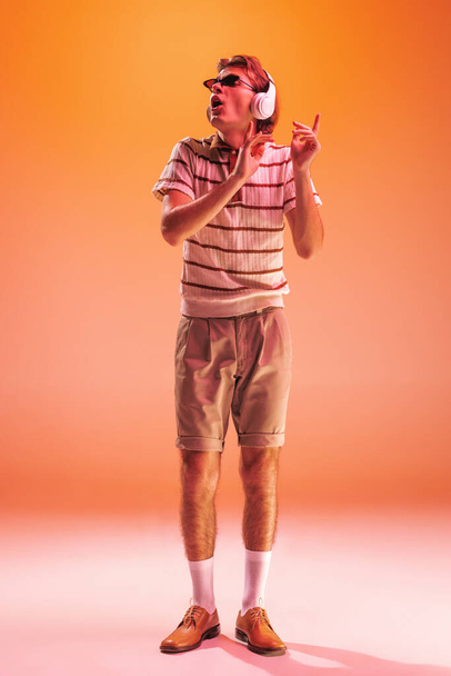 Retrato de jovem ouvindo música em fones de ouvido, dançando isolado sobre fundo estúdio laranja em luz de néon. Está a gostar da música. Conceito de juventude, moda, estilo de vida, emoções. Espaço de cópia para anúncio - Foto, Imagem