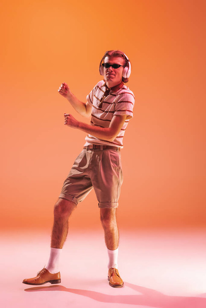 Πορτρέτο του νεαρού συναισθηματικού άνδρα που ακούει μουσική με ακουστικά, χορεύει απομονωμένος πάνω από πορτοκαλί φόντο στούντιο σε νέον φως. Έννοια της νιότης, της μόδας, του τρόπου ζωής, συναισθήματα. Αντιγραφή χώρου για διαφήμιση - Φωτογραφία, εικόνα