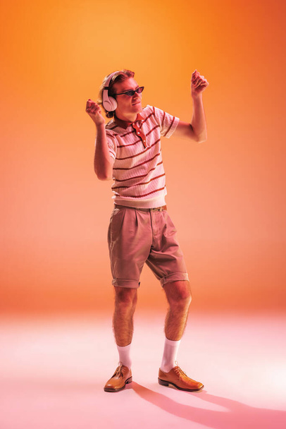 Portrait of young man listening to music in headphones, dancing isolated over orange studio background in neon light. Looks cheerful. Concept of youth, fashion, lifestyle, emotions. Copy space for ad - Foto, imagen