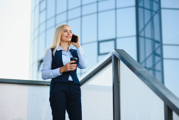Business Woman With Phone Near Office. Portrait Of Beautiful Smiling Female In Fashion Office Clothes Talking On Phone While Standing Outdoors. Phone Communication. High Quality Image - Foto, immagini